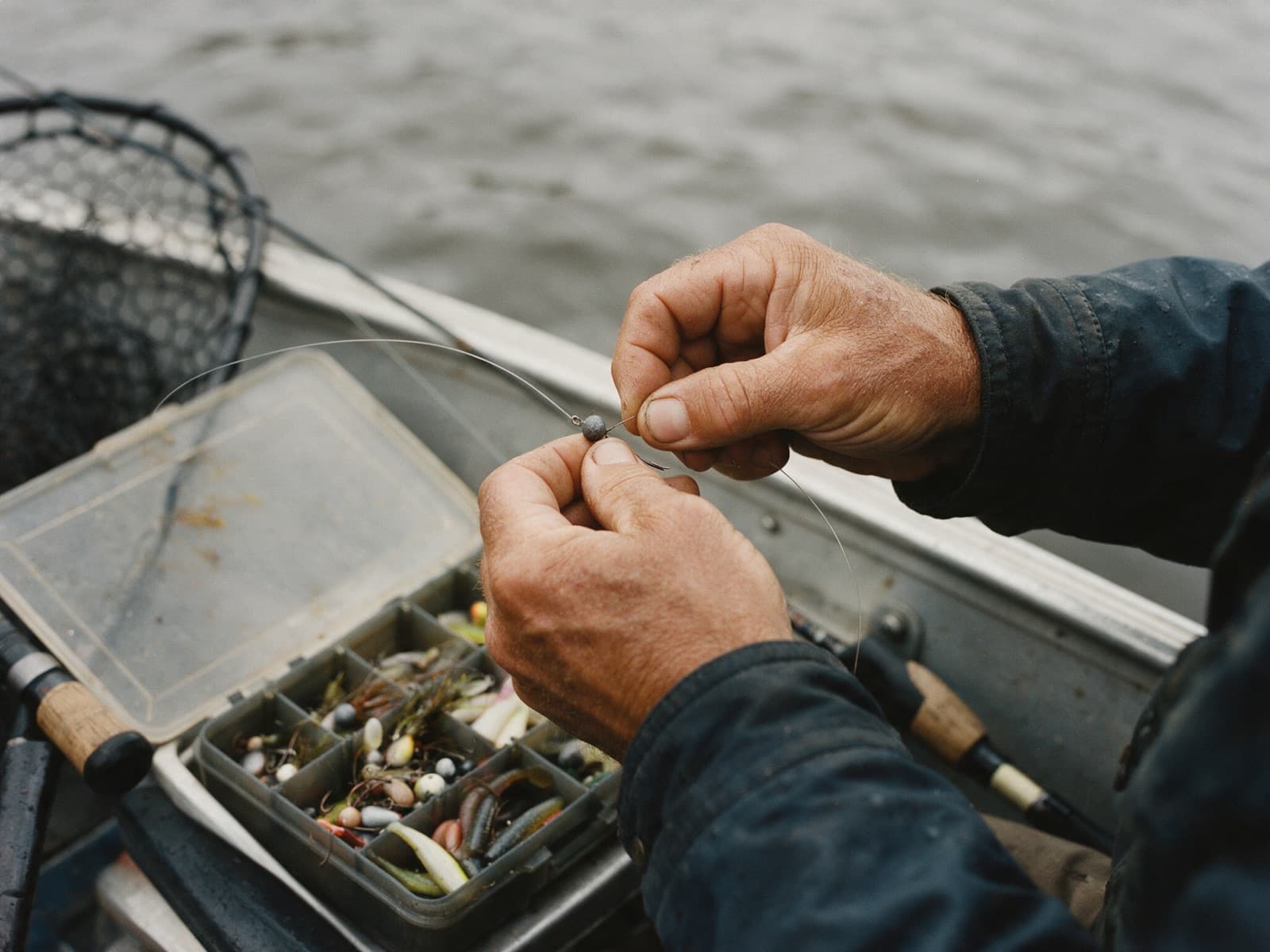 A guide's hands tying a jig onto line above an open tackle tray on a boat gunwale.